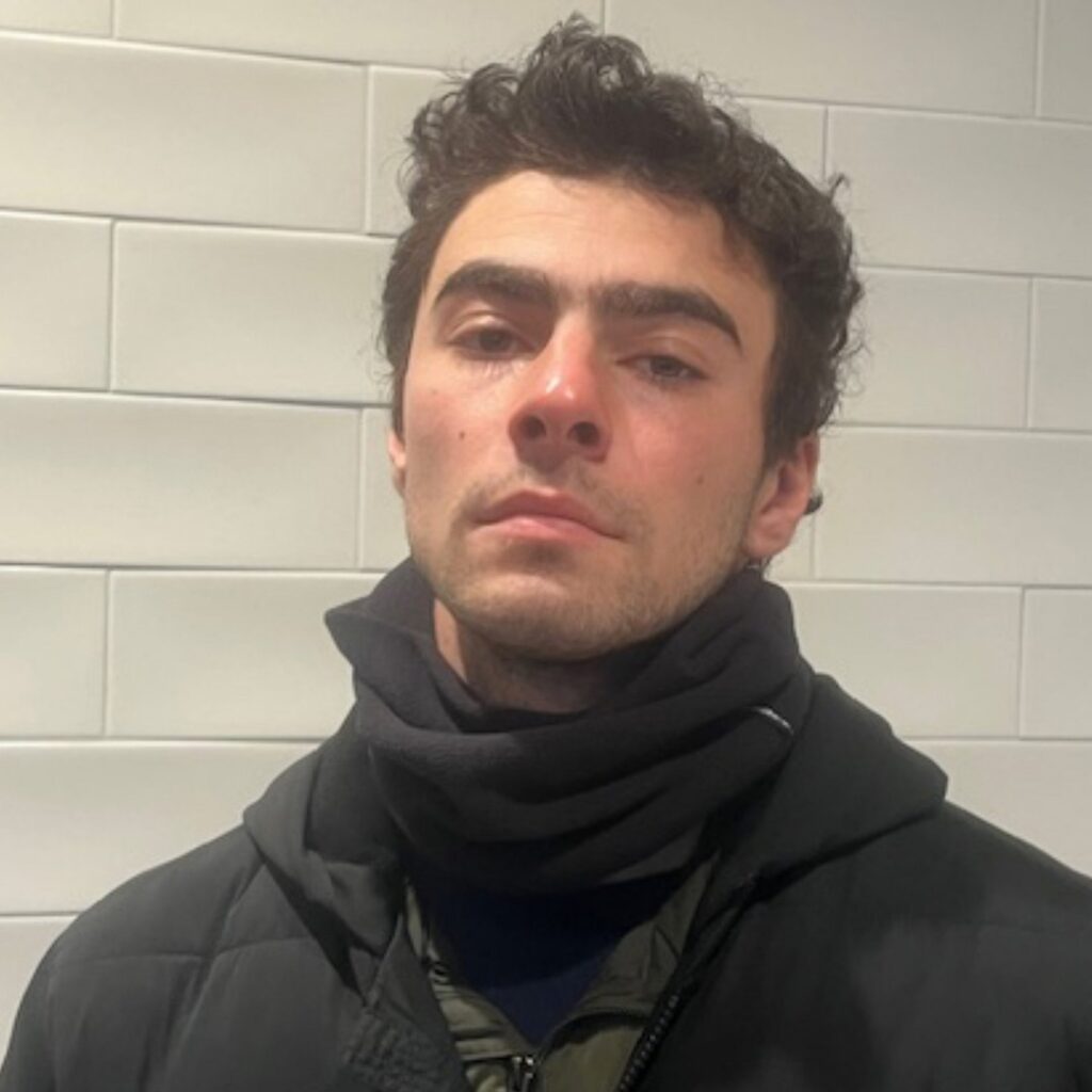 a police photo of Luigi Mangione wearing a black scarf and layers of dark jackets, with messy dark hair and bedroom eyes against a white brick wall on the day he was arrested